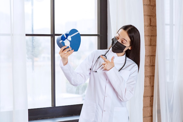 Médecin avec stéthoscope et masque noir tenant une boîte-cadeau en forme de coeur bleu.