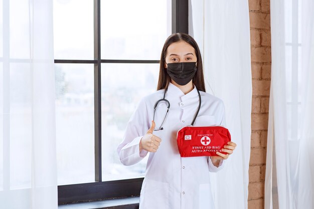 Médecin avec stéthoscope et masque noir tenant une trousse de premiers soins rouge et montrant le pouce vers le haut.