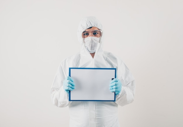 Un médecin tenant un tableau blanc sur fond clair dans des gants médicaux et une combinaison de protection. coronavirus