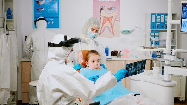 Médecin en tenue de protection discutant de la radiographie de la dent avec la mère d'un enfant patient expliquant le traitement à l'aide d'une tablette dans la pandémie de covisd-19 Équipe médicale portant un écran facial, une combinaison, un masque, des gants