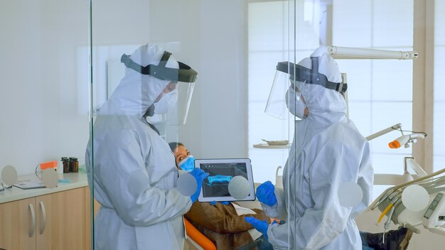 Médecins dentistes dans l'ensemble utilisant une tablette expliquant la radiographie dentaire dans un cabinet de stomatologie pendant le coronavirus. Homme portant un écran facial et un masque montrant à la radiographie de l'infirmière à l'aide d'un appareil numérique