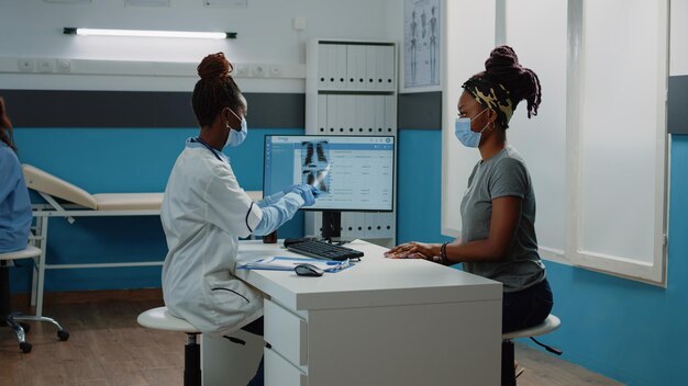 Medic tenant une radiographie pour examen avec le patient pendant la pandémie de coronavirus. Médecin expliquant les résultats de l'analyse aux rayons X et le diagnostic à la femme, portant des masques faciaux pour la protection.