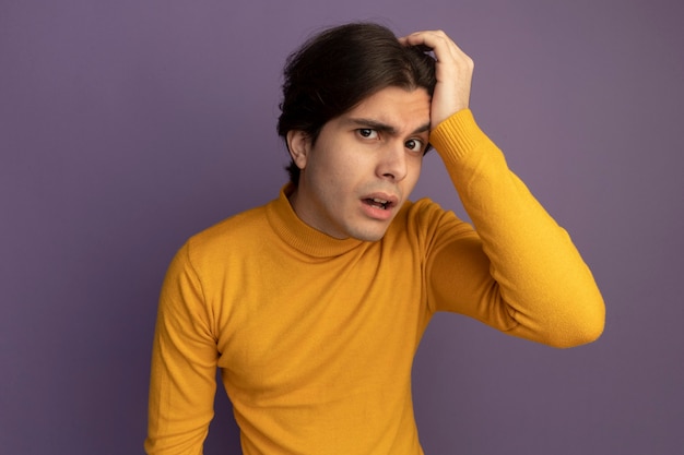 Méfiant jeune beau mec portant un pull à col roulé jaune mettant la main sur la tête isolé sur mur violet