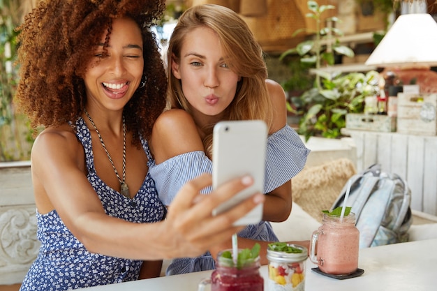 Les meilleures amies positives posent pour selfie, utilisent un téléphone portable moderne, s'assoient au restaurant, ont des expressions amusantes, partagent des photos sur les réseaux sociaux, connectées à Internet sans fil. Concept d'amitié