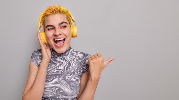 meloman fille aux cheveux jaunes teints points de maquillage lumineux pouce loin montre un espace vide écoute de la musique via des écouteurs sans fil sur gris