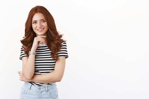 Membre de l'équipe féminine séduisante et créative en t-shirt rayé, touchant le menton et souriant heureux, entend un sujet intéressant, a l'air réfléchi et satisfait, choisit un produit en magasin