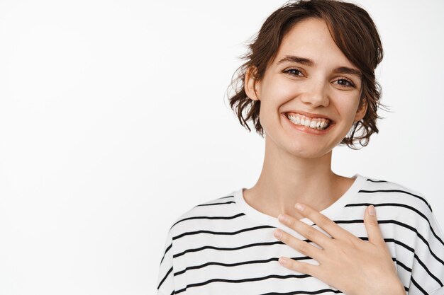 Merci. Une fille souriante et heureuse tient la main sur le cœur, l'air flattée, reconnaissante et satisfaite, debout sur un blanc