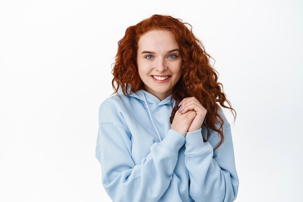 Merci. Souriante belle fille rousse avec une coiffure frisée, se tenant la main près de la poitrine, l'air reconnaissante, vous exprimer sa gratitude, debout sur blanc