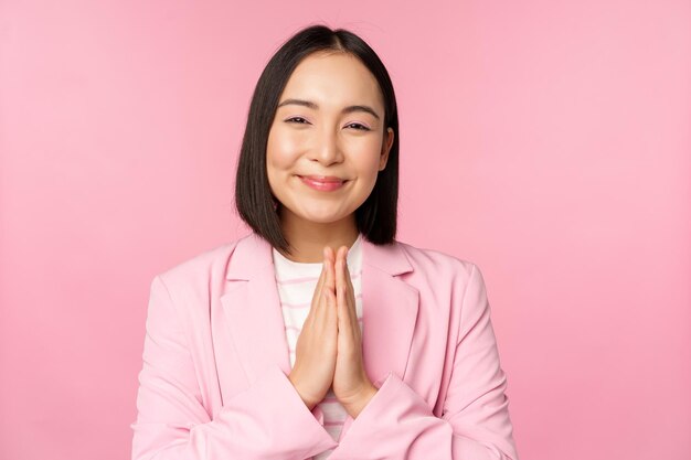 Merci Souriante vendeuse asiatique dame d'entreprise en costume remerciant main dans la main dans le sac geste de gratitude debout sur fond rose