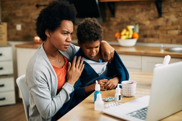 Une mère afro-américaine mesure la température de son fils tout en utilisant un ordinateur portable à la maison