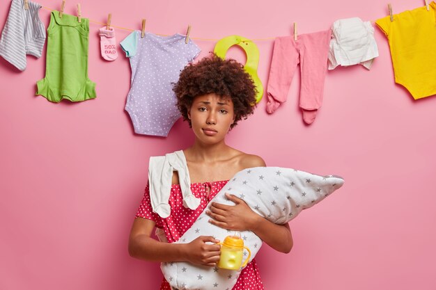 La mère aimante a l'air fatiguée après avoir allaité son bébé, tient le nouveau-né enveloppé dans une couverture, une bouteille de lait, pose contre un mur rose avec des vêtements d'enfant lavés suspendus à une corde. Concept de maternité