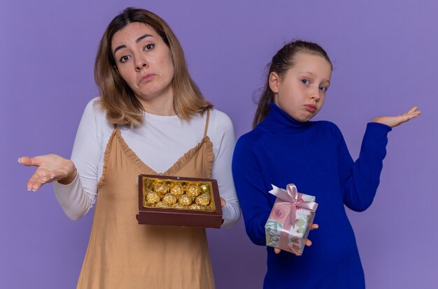 Mère aimante avec boîte de chocolats et fille tenant présent à lever les bras confus célébrant la journée internationale de la femme debout sur le mur violet