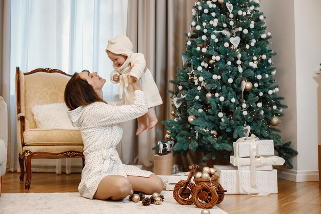 Mère Brune Et Petite Fille Assise Près De L'arbre De Noël Filles Portant Une Robe De Chambre Blanche Femme Tenant Sa Fille En L'air Photo Premium
