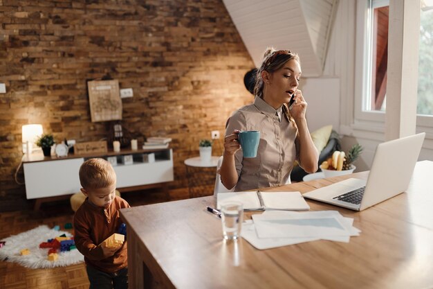 Mère célibataire parlant au téléphone en utilisant un ordinateur et en étant avec son fils à la maison