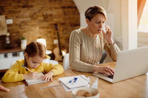 Mère choquée parlant au téléphone tout en lisant des e-mails problématiques sur un ordinateur portable à la maison