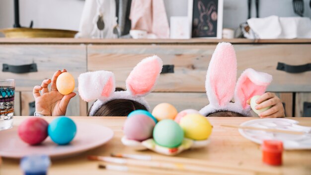 Mère avec un enfant dans des oreilles de lapin se cachant derrière une table avec des oeufs colorés