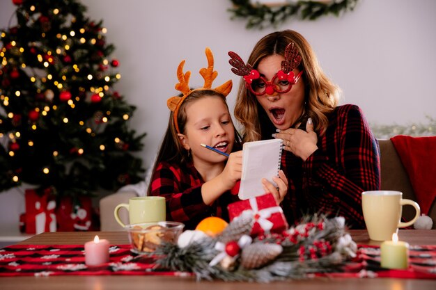Mère excitée dans des verres de rennes à la recherche de cahier avec sa fille assise à table en profitant du temps de Noël à la maison