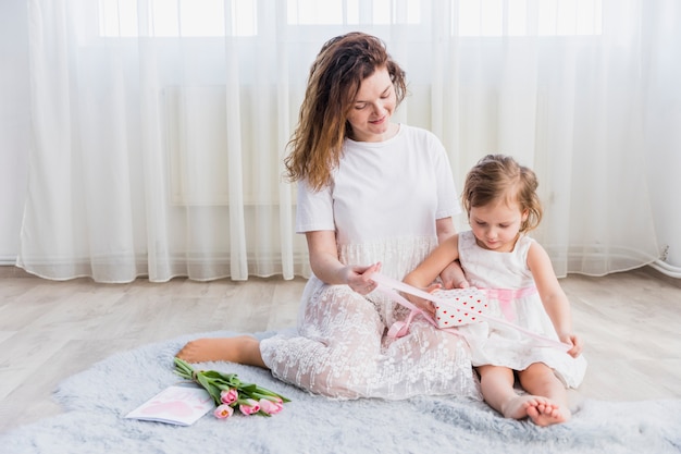 Mère et fille assise sur un tapis avec une boîte-cadeau; fleurs et carte de voeux