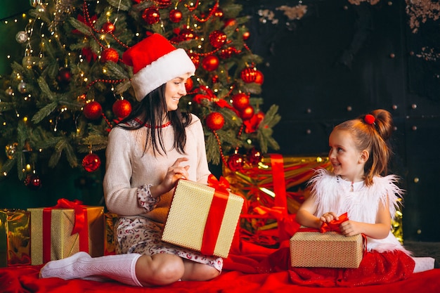 Mère avec fille par sapin de Noël déballant des cadeaux