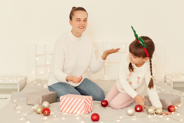 Mère et fille positives portant des chandails blancs de style décontracté assis sur le lit décorant avec des boules de Noël, femme avec bras levé et regardant la caméra avec le sourire.