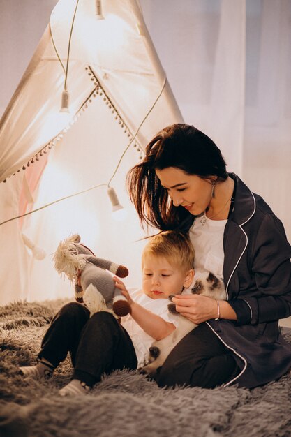 Mère avec fils assis dans une tente confortable avec des lumières à la maison à Noël