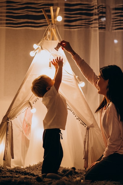 Mère avec fils assis dans une tente confortable avec des lumières à la maison à Noël