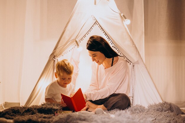 Mère avec fils assis dans une tente confortable avec des lumières à la maison à Noël