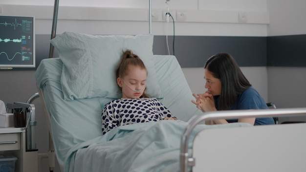 Mère inquiète assise à côté de sa petite fille priant pendant son sommeil après une opération chirurgicale pour une maladie en attente d'une expertise médicale dans la salle d'hôpital. Enfant malade portant un tube nasal pendant le traitement médicamenteux