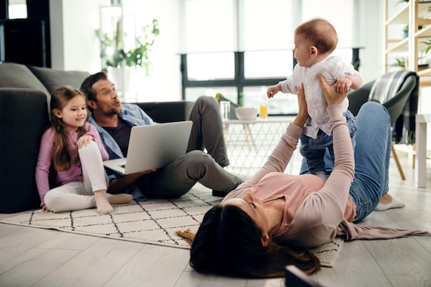 Mère ludique tenant son bébé en position allongée sur le sol dans le salon Père et fille utilisent un ordinateur portable