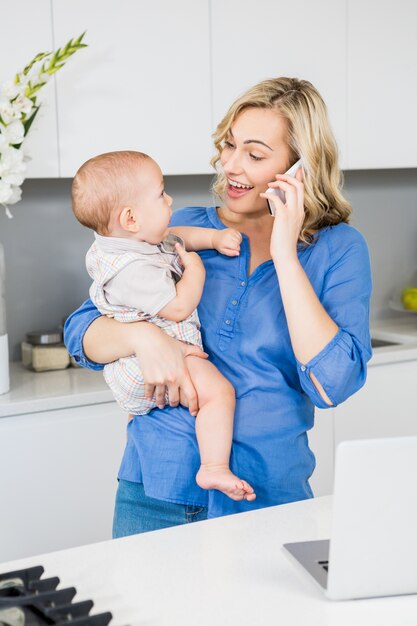 Mère parler sur téléphone mobile tout en tenant son petit garçon dans la cuisine