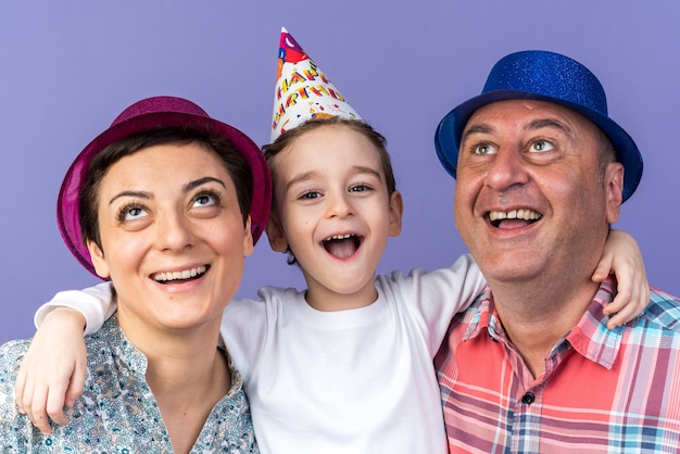 mère et père joyeux avec des chapeaux de fête levant debout avec leur fils isolé sur un mur violet avec espace de copie