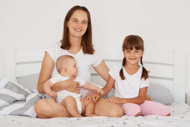 Mère avec ses jolies petites filles assises sur le lit, profitant ensemble de leur temps libre, regardant la caméra avec des expressions faciales heureuses, une maternité heureuse, la parentalité.