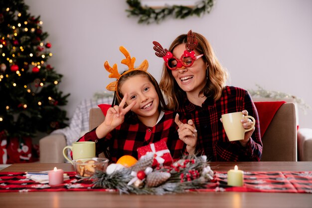 mère souriante tenant une tasse et sa fille gesticulant le signe de la victoire assis à table profitant de la période de Noël à la maison