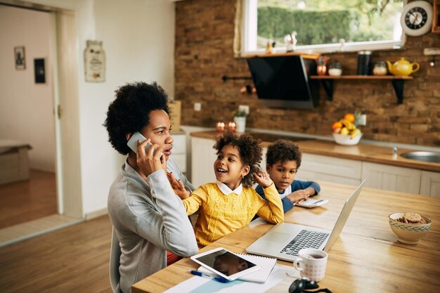 Mère travailleuse noire parlant au téléphone tout en étant à la maison avec ses enfants