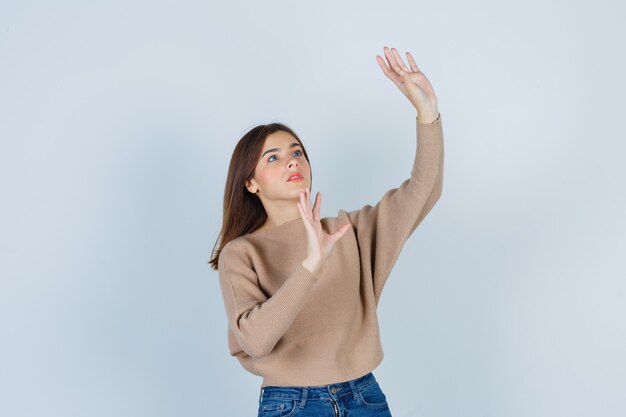 Merveilleuse Dame Levant Les Mains De Manière Protectrice En Pull, Jeans Et Ayant L'air Effrayée. Vue De Face.