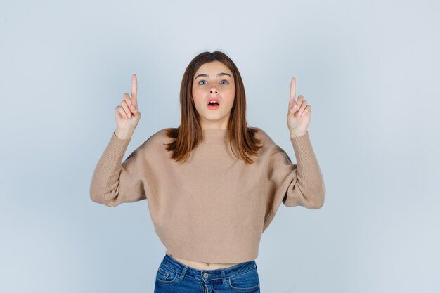 Merveilleuse dame pointant vers le haut et ouvrant la bouche dans un pull, un jean et l'air choquée. vue de face.