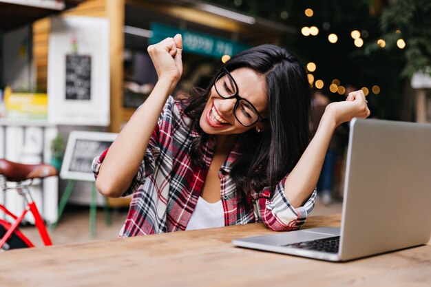 Merveilleuse étudiante assise dans un café avec ordinateur portable et riant. Indépendant agréable dans des verres exprimant le bonheur.