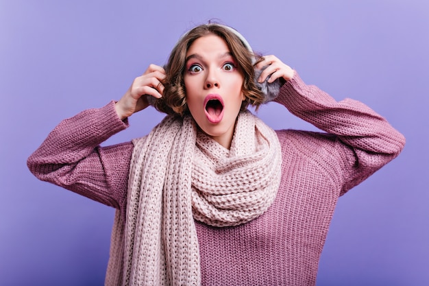 Merveilleuse femme brune avec de grands yeux touchant ses écouteurs en fourrure. choqué fille pâle en écharpe posant avec la bouche ouverte.