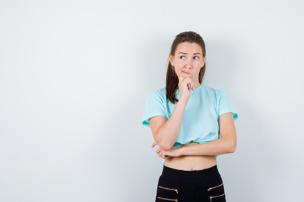 Merveilleuse femme en chemisier, pantalon gardant la main sur le menton tout en regardant de côté et l'air pensif, vue de face.