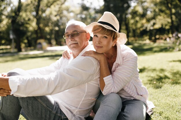 Merveilleuse femme avec une coiffure blonde en chapeau à la mode et chemise rose assis sur l'herbe avec un homme à la moustache et des vêtements blancs dans le parc.