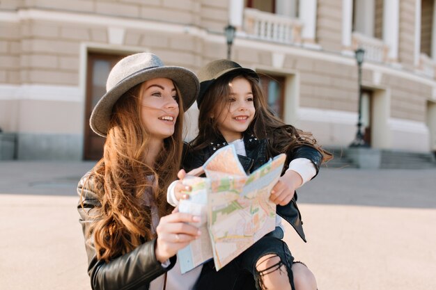 Merveilleuse femme frisée inspirée au chapeau tenant jolie fille et plan de la ville, à la recherche de suite. Portrait en plein air de deux filles voyageant autour d'un nouvel endroit et recherchant de beaux sites.