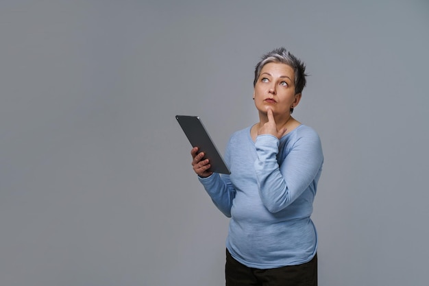 Merveilleuse femme mature des années 50 aux cheveux gris avec tablette numérique travaillant ou vérifiant sur les réseaux sociaux Jolie femme en blouse bleue isolée sur blanc Personnes âgées et technologies Image tonique