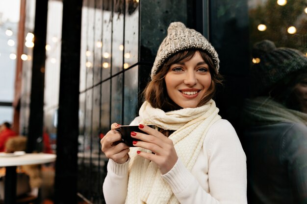 Merveilleuse femme mignonne avec un grand sourire portant un bonnet tricoté et un pull tenant une tasse de café et profiter d'une pause-café à l'extérieur. Photo de haute qualité