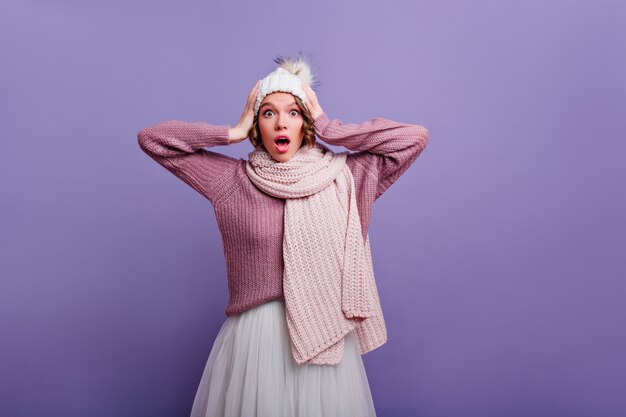 Merveilleuse femme surprise avec une écharpe tricotée touchant sa tête. Portrait intérieur d'une fille caucasienne choquée en chapeau et jupe blanche.