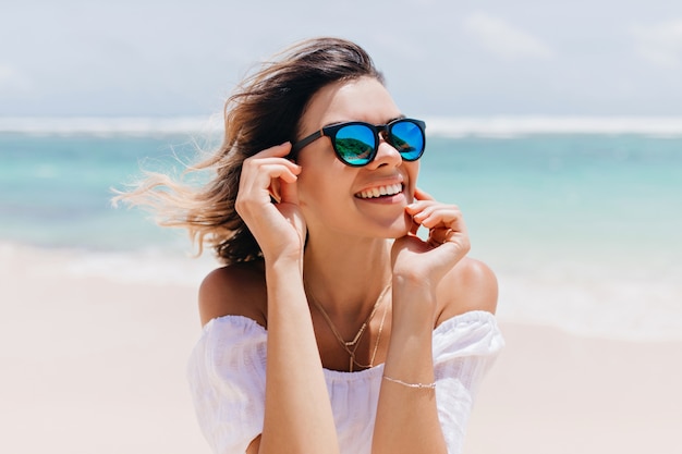 Merveilleuse femme en tenue blanche et lunettes scintillantes posant avec l'expression du visage heureux en chaude journée d'été Agréable femme caucasienne debout près de l'océan sur le ciel