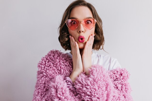 merveilleuse fille caucasienne exprimant des émotions surprises sur un mur de lumière. Photo d'une femme étonnée extatique en manteau de fourrure rose et lunettes de soleil.