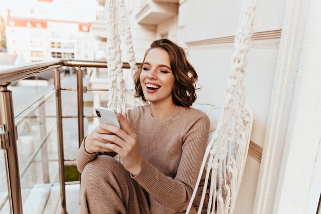 Merveilleuse fille lisant un message téléphonique assis à la terrasse. Photo de femme mignonne excitée posant avec smartphone au balcon.