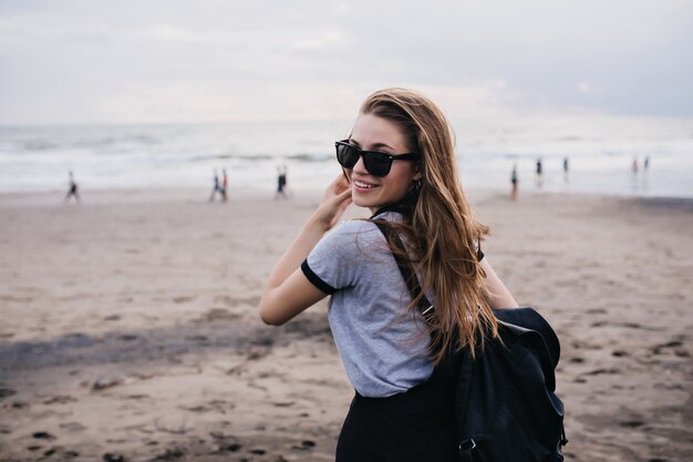 Merveilleuse fille en lunettes de soleil noires regardant par-dessus l'épaule en se tenant debout sur la plage de sable Photo de beau modèle féminin avec blackpack posant sur la nature.