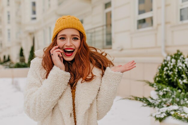 Merveilleuse fille rousse parlant au téléphone en journée d'hiver Photo extérieure d'une adorable jeune femme en manteau