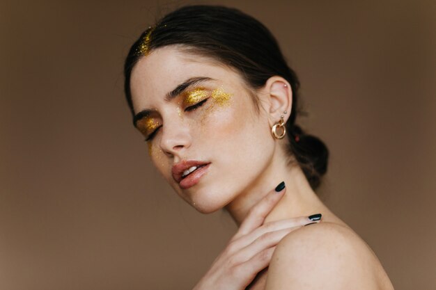 Merveilleuse jeune femme avec une élégante boucle d'oreille dorée posant sur un mur sombre. Femme brune extatique avec maquillage de fête debout avec les yeux fermés.